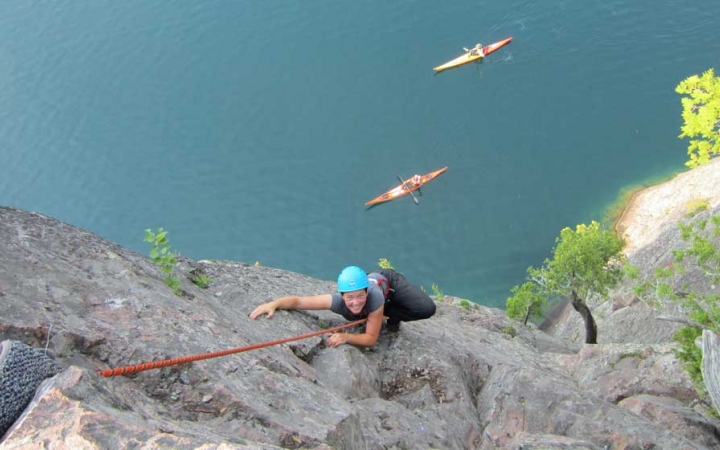 A person wearing safety gear and secured by ropes pauses their climb to look up at the camera. Below them, two kayaks rest on vast blue water. 